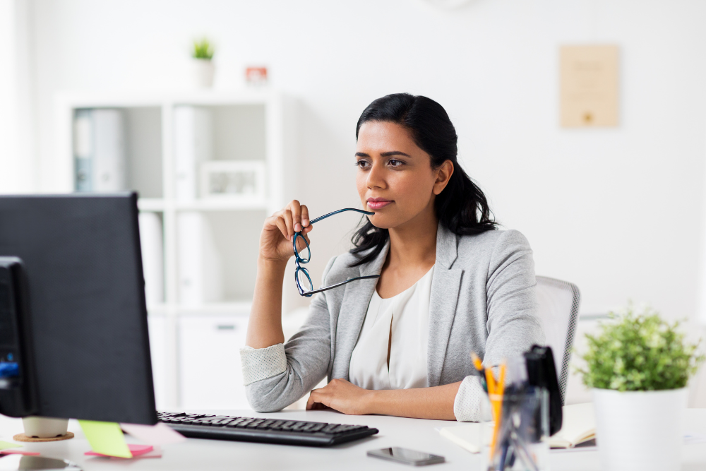 A person sits at a desk with a computer, holding glasses and looking off to the side thoughtfully.