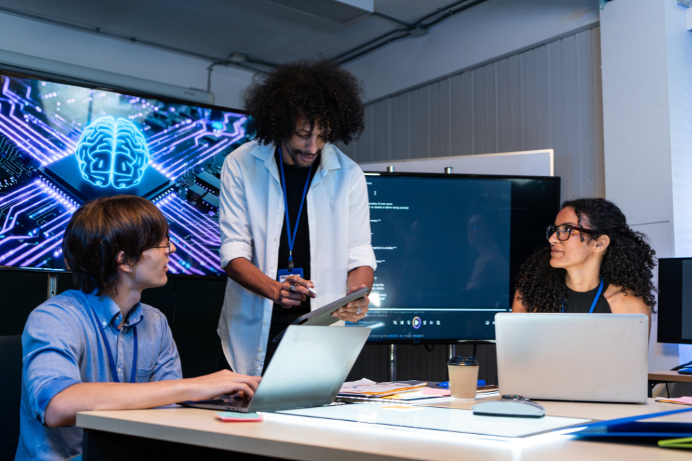 Three people are gathered around a table in a dimly lit room, with a large screen displaying a blue, circuit-board-like brain graphic behind them. 
