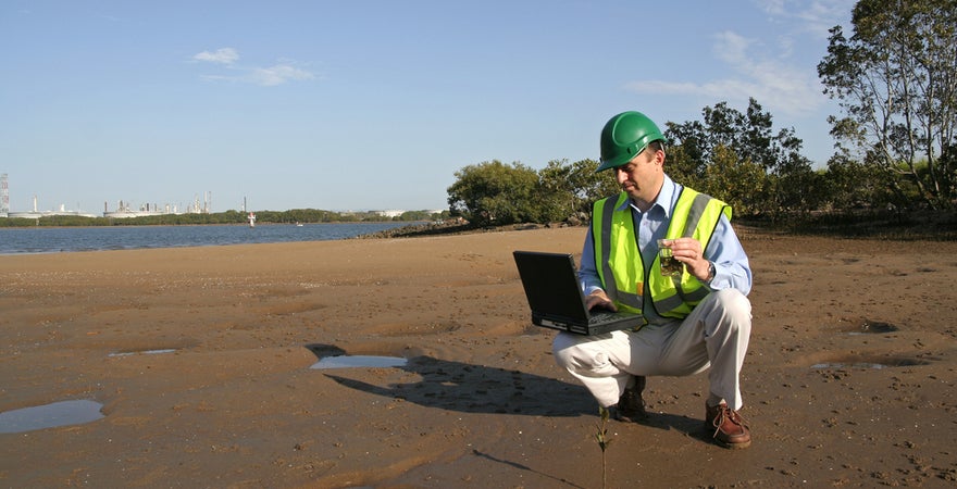 An environmental scientist stood on mudflats analysing data on his laptop.