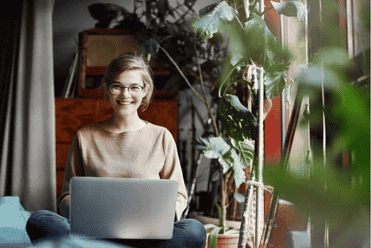 A smiling woman sitting near a window with a laptop surrounded by greenery