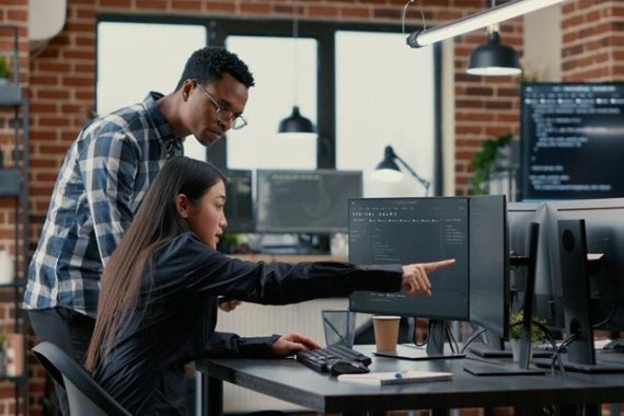 Two people looking intently at a computer screen in an office setting, the woman sitting and pointing while man stands looking at the same screen. 
