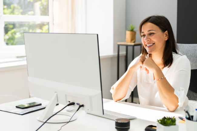 A smiling person sits at a desk in front of a computer, with their hands clasped in front of their chin. Sunlight streams in from a window behind the monitor.