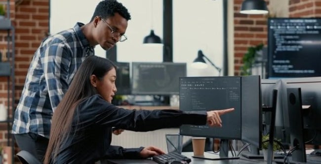 Two people looking intently at a computer screen in an office setting, the woman sitting and pointing while man stands looking at the same screen.