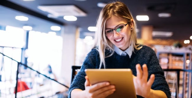 Woman with long blonde hair uses a tablet in a modern cafe or workspace, illustrating participation and advancement of women in the finance sector.