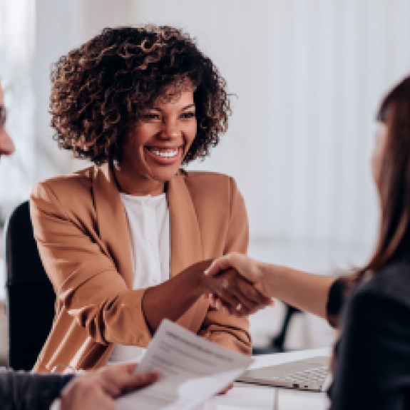 Two women shaking hands in a business meeting while male colleague smiles on the left.