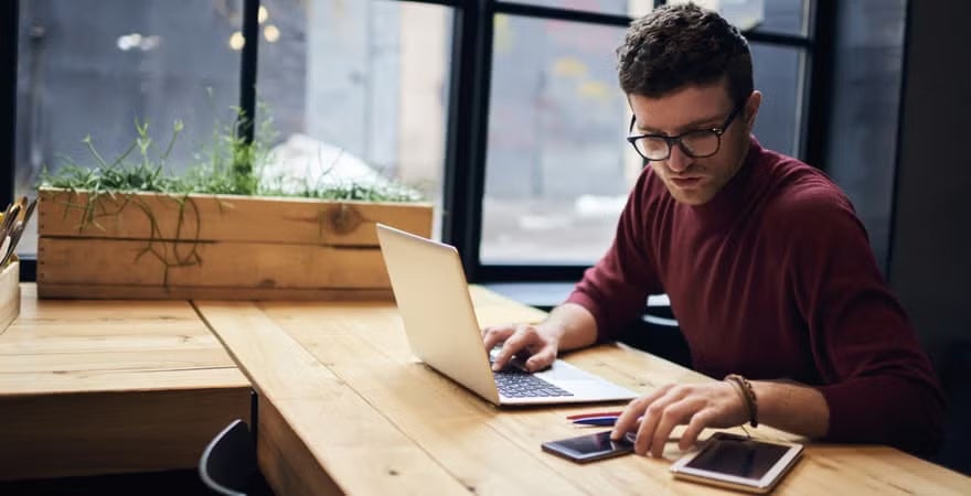 A man in a maroon shirt works at a laptop in a modern workspace with large windows, a wooden planter, and digital devices on the table.