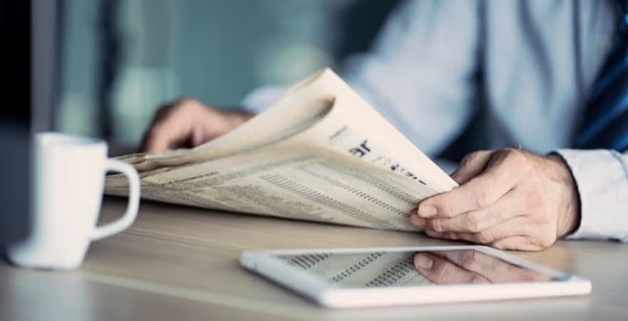 A businessman reading a newspaper; coffee and tablet in the foreground.
