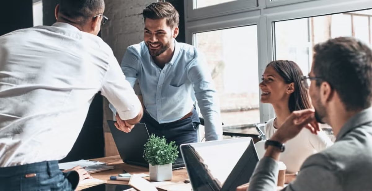 Group of professionals in a meeting room, one person stands and shakes hands with a colleague, while others smile and interact around laptops, suggesting leadership and teamwork