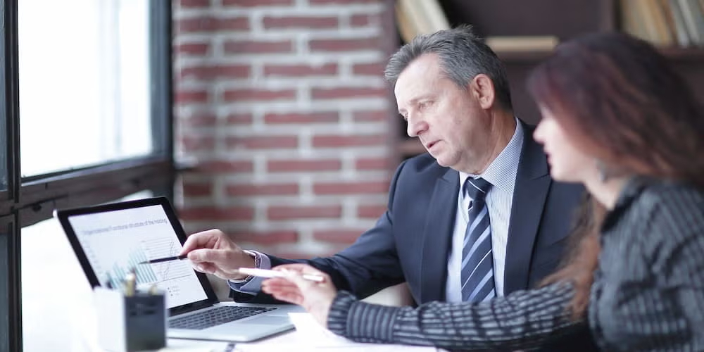 Two professionals, one man in a suit and one woman, discuss charts on a laptop in a modern office, focusing on analysis and strategy.