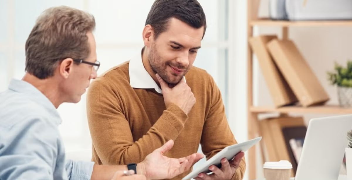 Two men in discussion at a desk, one holding a tablet and the other gesturing, with bookshelves and a laptop in the background, illustrating academic or educational consultation.